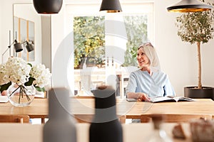 Senior Woman Relaxing At Home Reading Magazine Sitting At Dining Room Table