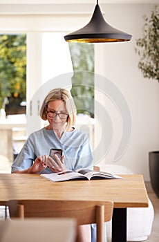 Senior Woman Relaxing With Magazine At Home Looking At Mobile Phone Sitting At Dining Room Table