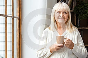 Senior woman resting at home standing near the window drinking coffee elderly lifestyle