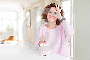 Senior woman saving money putting a coin inside piggy bank with happy face smiling doing ok sign with hand on eye looking through