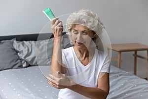 Senior woman sit on bed apply nourishing cream on hands