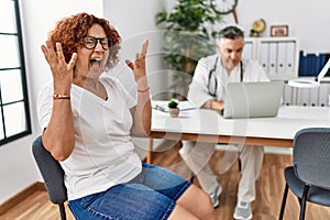 Senior woman sitting at doctor appointment celebrating mad and crazy for success with arms raised and closed eyes screaming