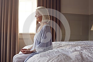 Senior Woman Sitting On Edge Of Bed At Home Meditating During Lockdown For Covid-19