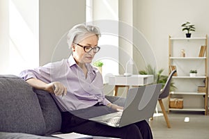 Senior woman sitting with laptop on couch at home and having video call or watching webinar