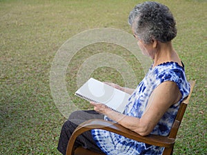 Senior woman sitting reading a book at the chair in the garden. Space for text. Concept of aged people and relaxation