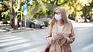 Senior woman with smartphone crossing street outdoors in city or town, coronavirus concept.