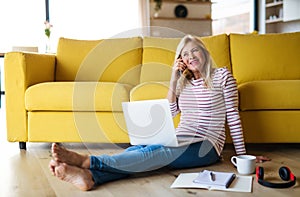 Senior woman with smartphone and laptop indoors in home office, working.