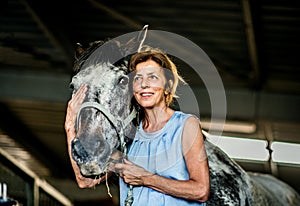 A senior woman standing close to a horse in a stable, holding it.