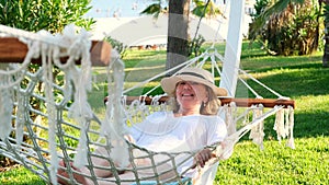senior woman in straw hat smiling happy relaxing on a hammock enjoying the fresh air on the terrace around palm trees