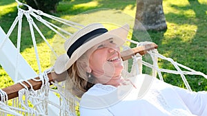 senior woman in straw hat smiling happy relaxing on a hammock enjoying the fresh air on the terrace around palm trees