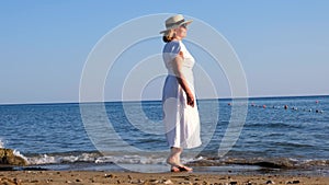 senior woman in sunglasses in a straw hat and white dress walks along the blue sea coast on a sunny summer day, enjoying