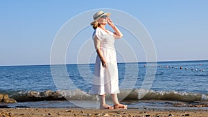 senior woman in sunglasses in a straw hat and white dress walks along the blue sea coast on a sunny summer day, enjoying