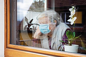 Senior woman with surgical mask sitting on a window at home, coronavirus and covid-19 provisions photo