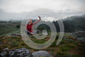 Senior woman taking selfie when doing breathing exercise in nature on early morning with fog and mountains in background