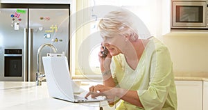 Senior woman talking on mobile phone while using laptop in kitchen