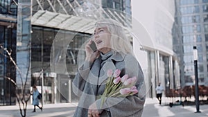 A senior woman talking by phone holding tulips outdoors