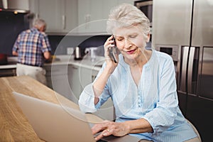 Senior woman talking on phone while using laptop and man working in kitchen