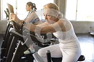 Senior woman tired exercising spinning bike in fitness gym.