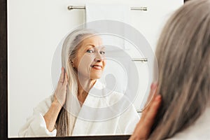 Senior woman touching her long grey hair and looking at a mirror in bathroom