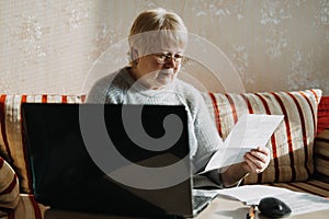 Senior woman using laptop and holding documents at home. Focused mature aged female using computer working with finance