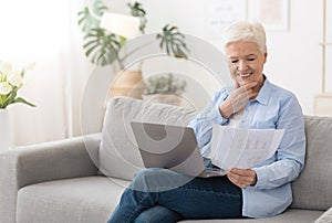 Senior woman using laptop and reading documents while working remotely at home