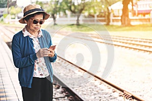 Senior woman using a mobile phone while sitting on bench in train station. Asian grandmother standing and using a smart phone