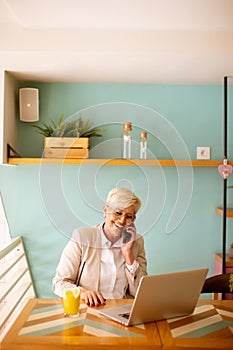 Senior woman using mobile phone while working on laptop and drinking fresh orange juice in the cafe