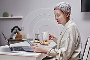 Senior woman using smartphone and working while enjoying breakfast in morning