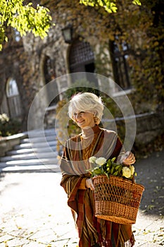 Senior woman walking with basket full of flowers and groceries in autumn park