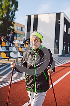 Senior woman walking with walking poles in stadium on a red rubber cover. Elderly woman 88 years old doing Nordic walking
