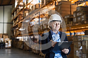 Senior woman warehouse manager or supervisor with white helmet and clipboard.