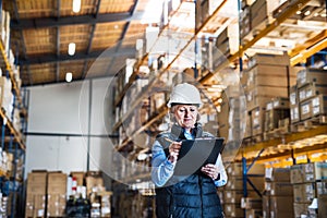 Senior woman warehouse manager or supervisor with white helmet and clipboard.