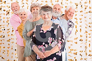 Senior woman wearing elegant blouse standing in front of group of senior happy fiends looking from behind her