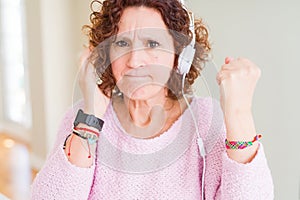 Senior woman wearing headphones listening to music annoyed and frustrated shouting with anger, crazy and yelling with raised hand,