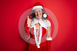 Senior woman wearing Santa Claus costume holding christmas gift over isolated red background with a happy face standing and