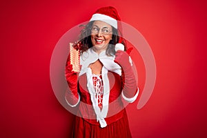 Senior woman wearing Santa Claus costume holding christmas gift over isolated red background pointing and showing with thumb up to