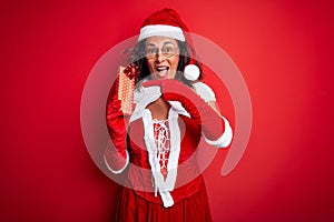 Senior woman wearing Santa Claus costume holding christmas gift over isolated red background very happy pointing with hand and