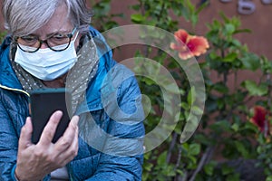 A senior woman wearing a surgical mask due to coronavirus sitting in a blossom garden looking at her smartphone. Old retiree using