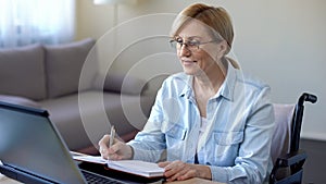 Senior woman in wheelchair studying online and making notes, video lessons