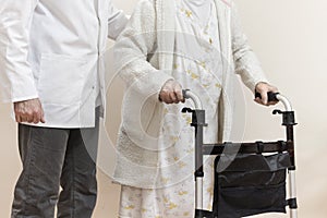 Senior woman in a white bathrobe and nightgown goes with a rehabilitation walker. The nurse supports and protects the old wom