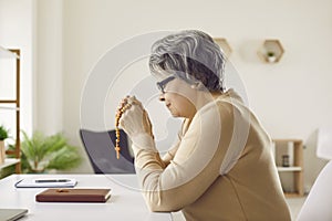 Senior woman who belongs to Catholic Christian Church sitting at desk and praying