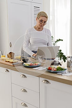 Senior woman working at laptop in kitchen. Looks cooking recipes online.