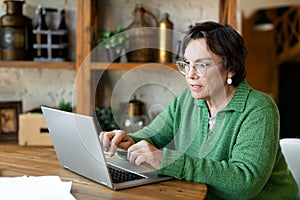 Senior woman works in front of a laptop monitor with papers at home. Home Office. She goes through tax returns and