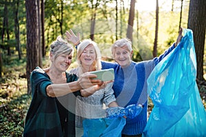 Senior women friends picking up litter outdoors in forest, taking selfie.