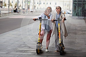 Senior women with gray hair enjoying ride together on electric scooters outdoors on streets. Elderly female friends