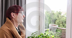 Senior women looking out of window at home. Depressed old aged woman looking outside of a window during quarantine and lockdown