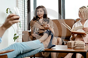 Senior Women Spending Time Together At Drawing Room. Blonde Woman Reading Book. Her Friend Steetched Legs Above Coffee Table.