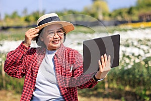 Senior women wearing plaid shirt and hat video call by tablet with daring of her in flower garden, elderly people tech and social