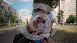 Senior 90-year-old woman with gray hair and deep wrinkles sits outdoors In assisted living facility on bench with small