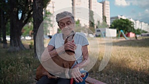 Senior 90-year-old woman with gray hair and deep wrinkles sits outdoors In assisted living facility on bench with small